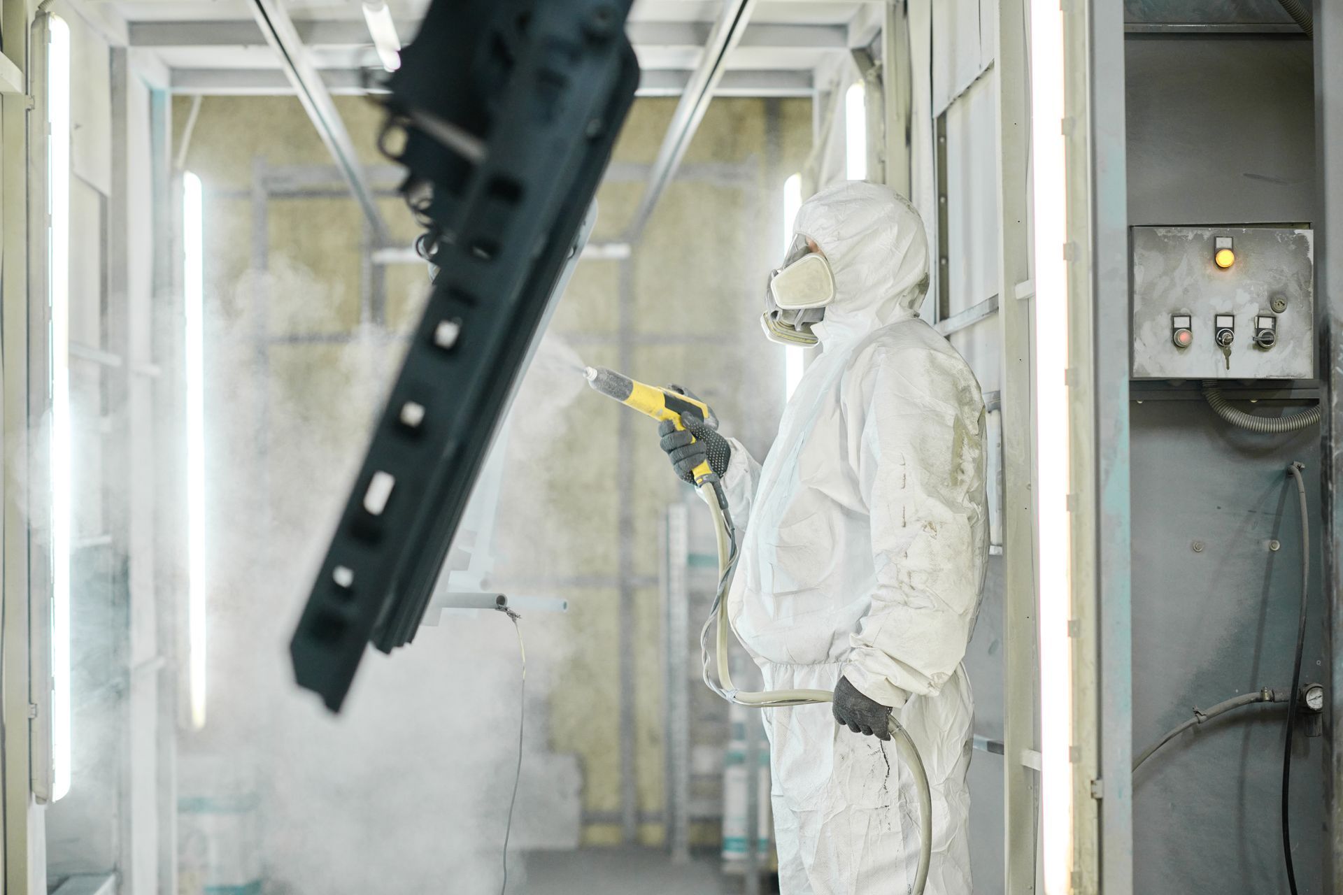 Person in protective suit powder coating a black metal object in an industrial setting.