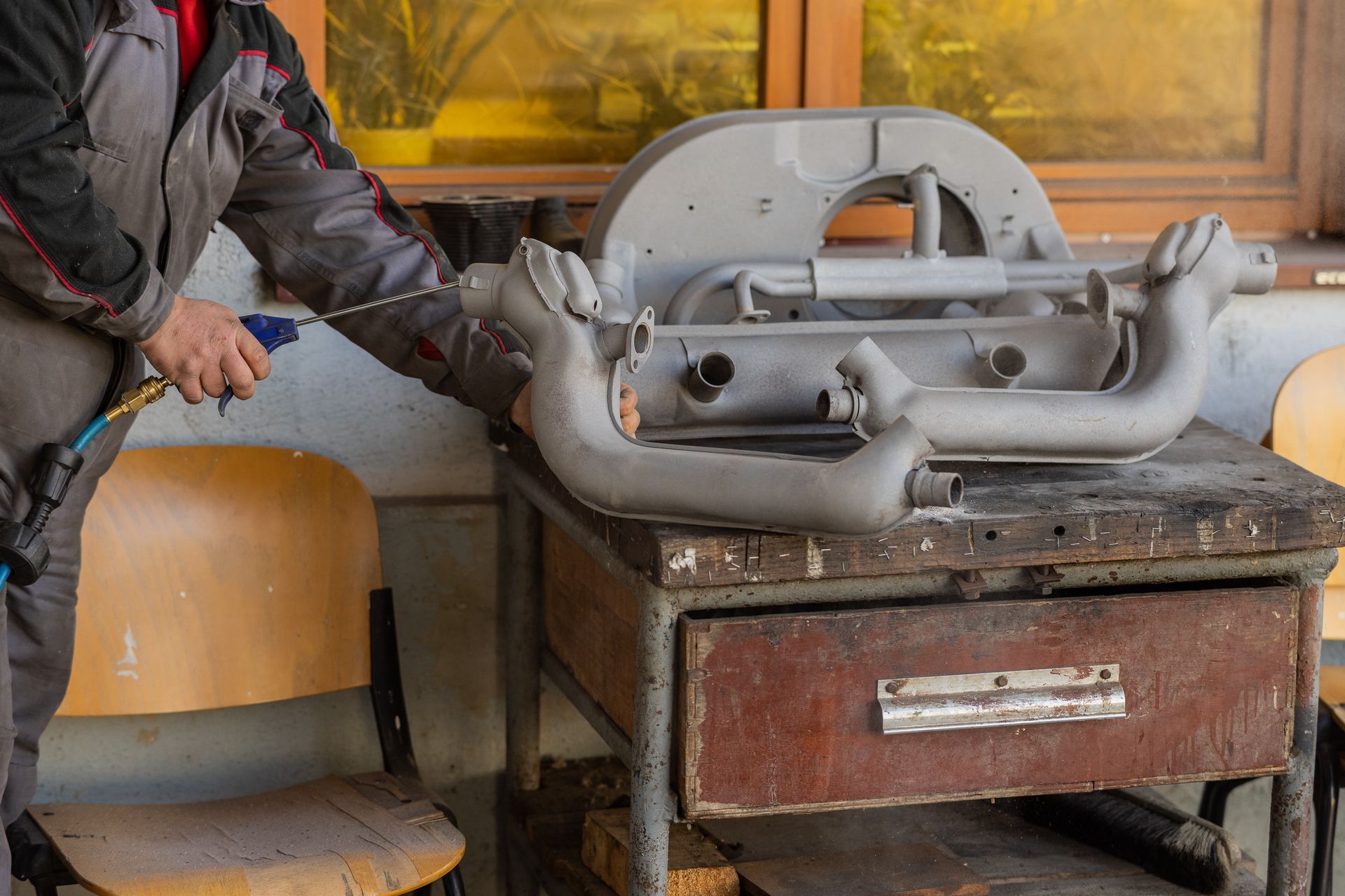 Person spraying a gray engine part on a workbench near a window.