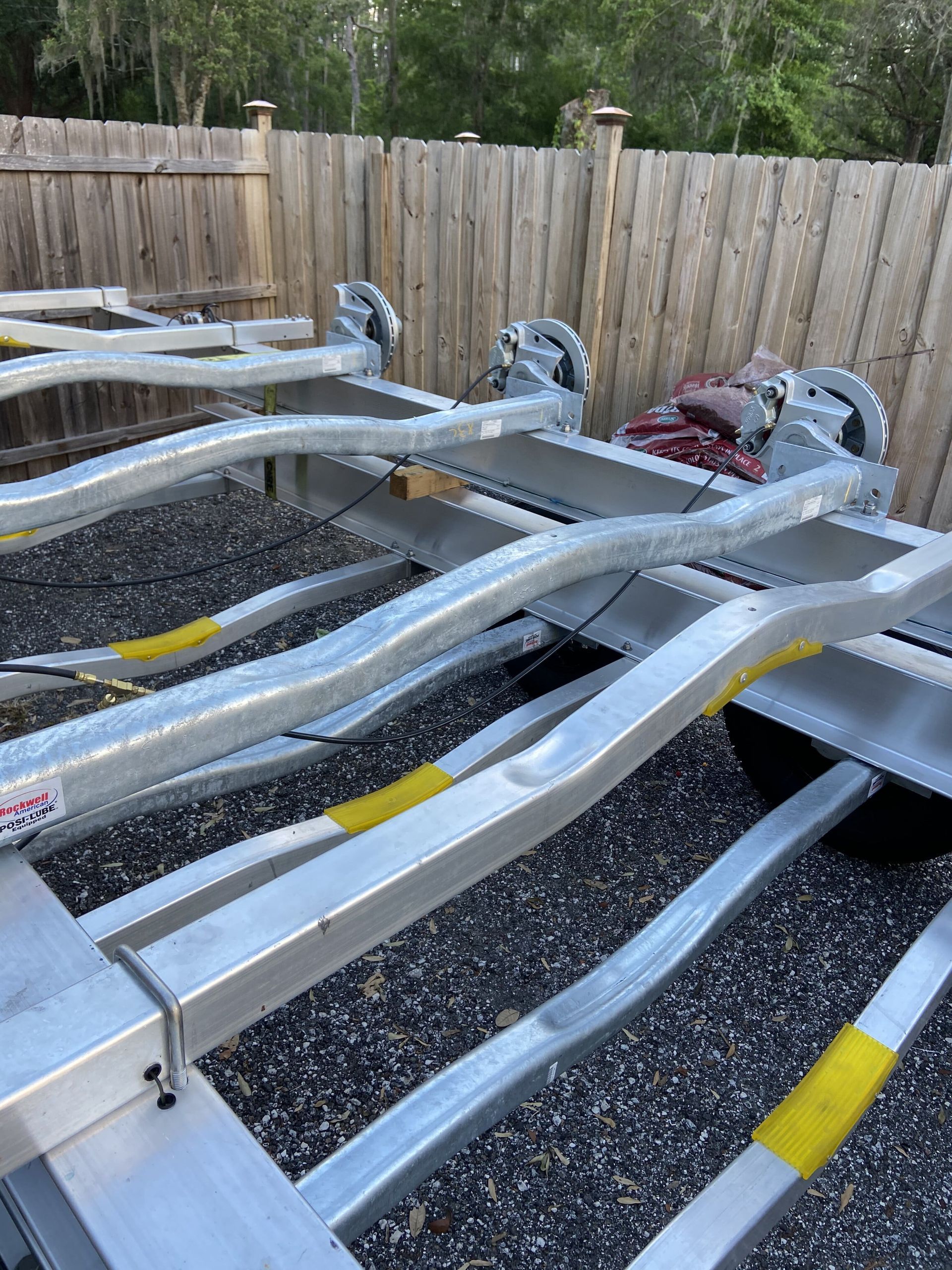 A boat trailer is sitting on gravel in front of a wooden fence.
