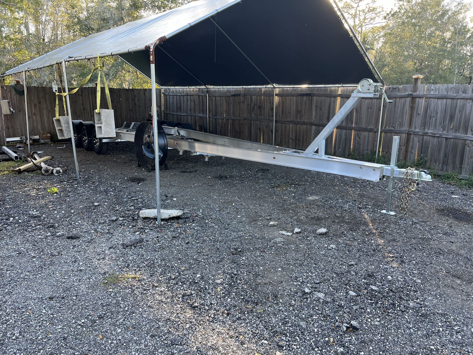 A boat trailer is parked under a canopy in a gravel lot.