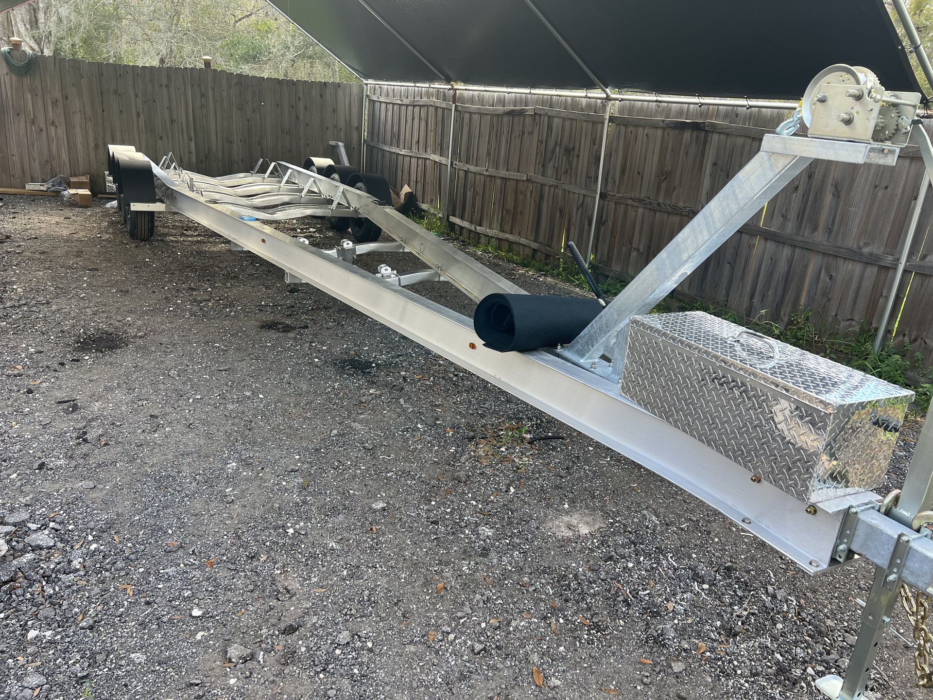 A boat trailer is parked in a gravel lot next to a wooden fence.