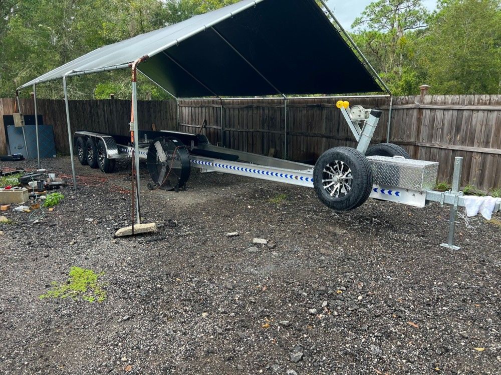 A boat trailer is parked under a canopy in a gravel lot.