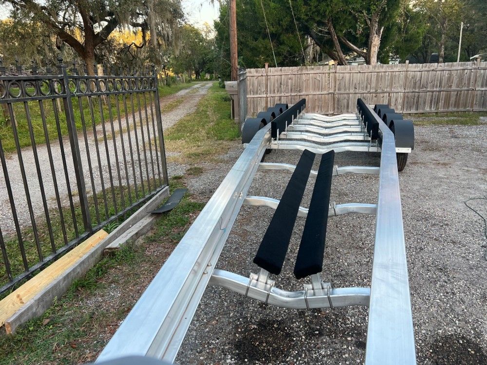 A boat trailer is parked in a gravel lot next to a fence.
