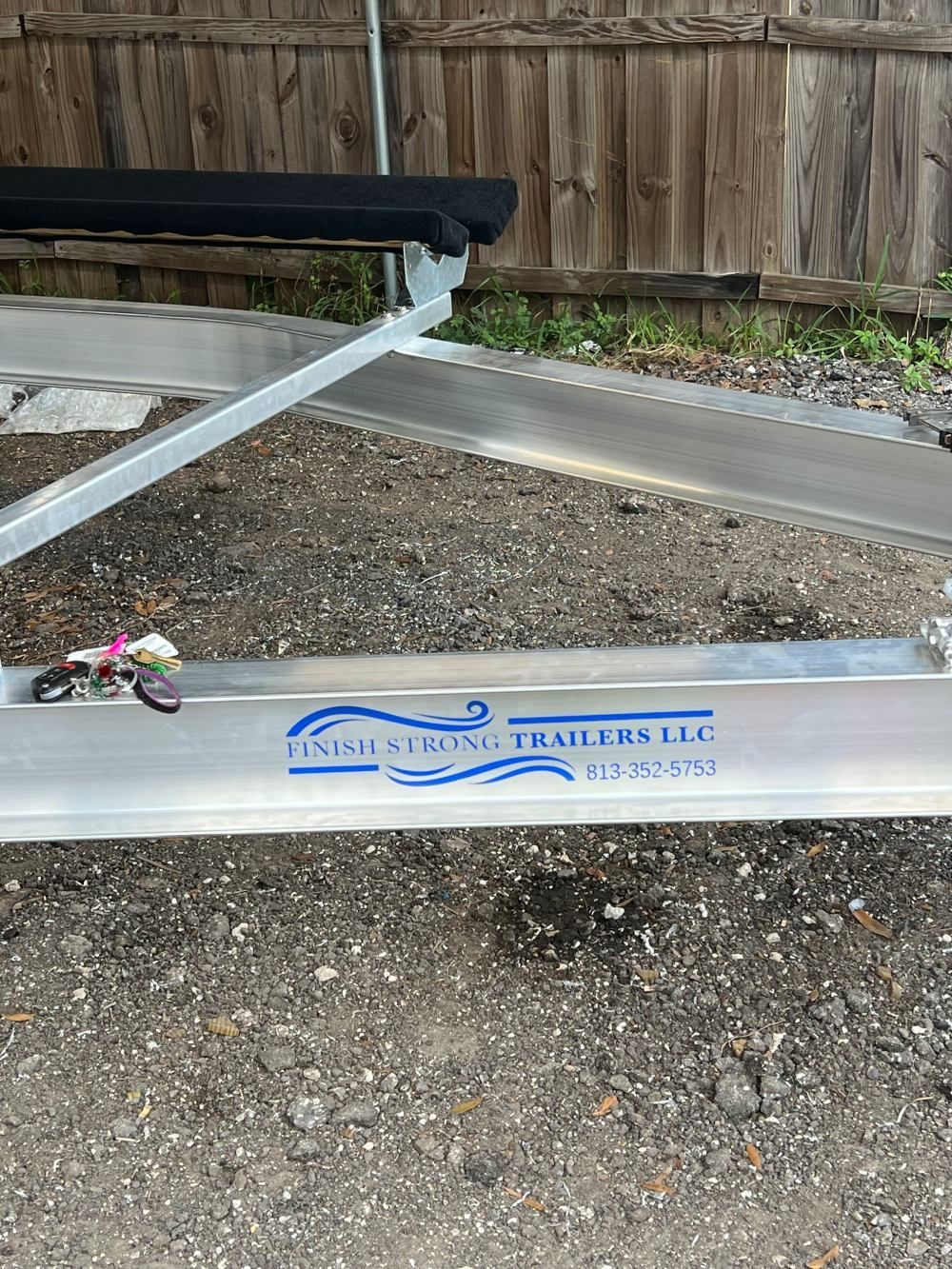 A boat trailer is parked in a gravel lot next to a wooden fence.