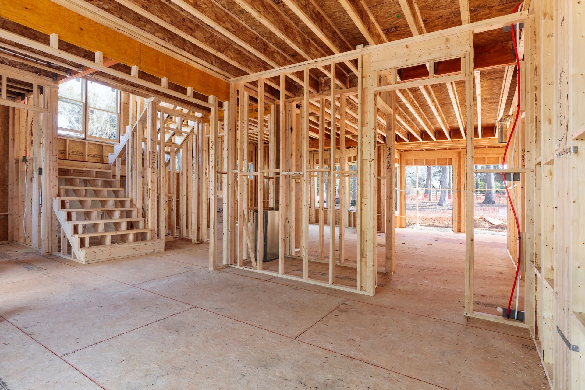 Interior view of a house under construction with exposed wooden framing, subflooring, and a partially built staircase.
