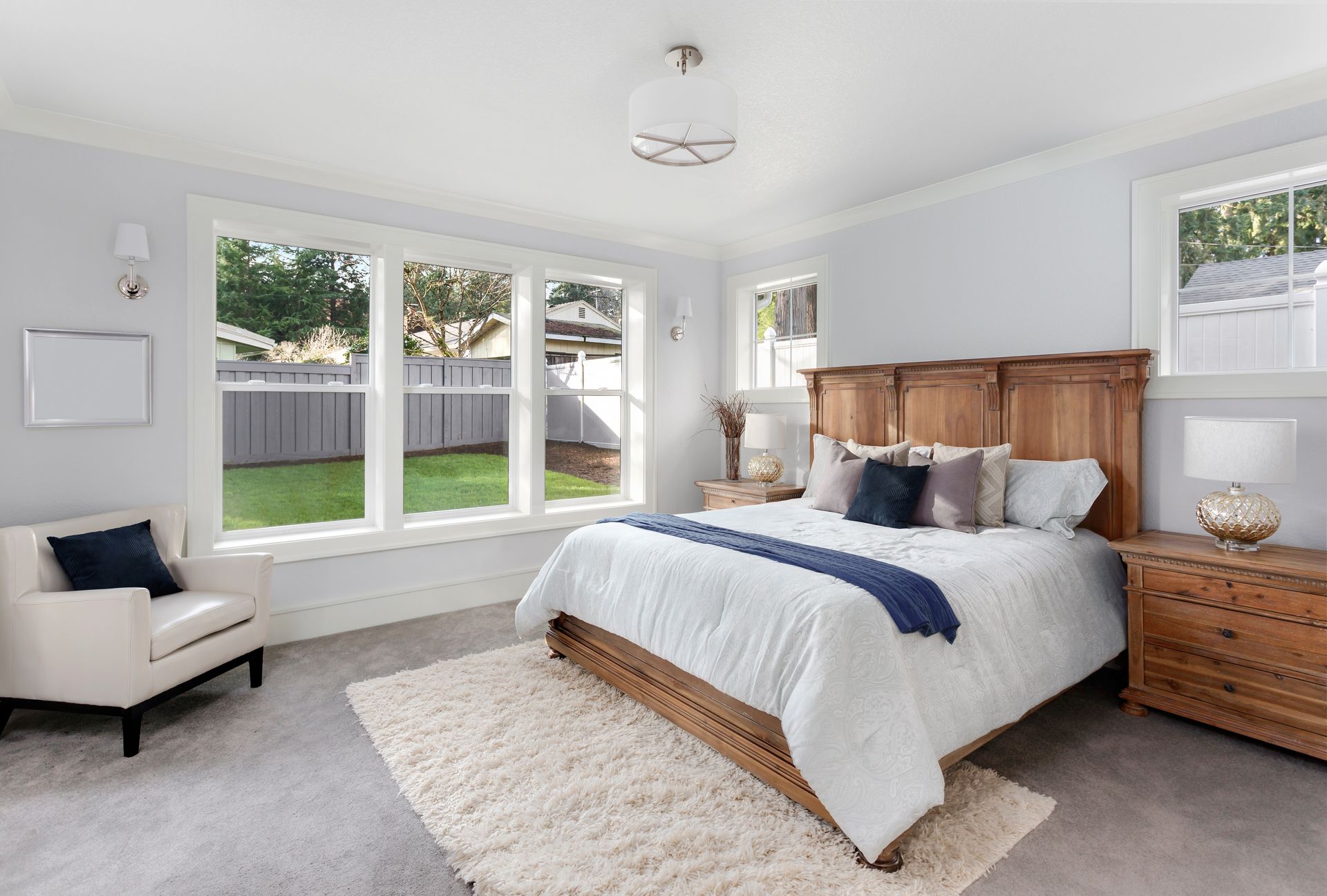 A bright, contemporary bedroom features a king bed with a wood headboard, a white armchair, and large windows with garden views.