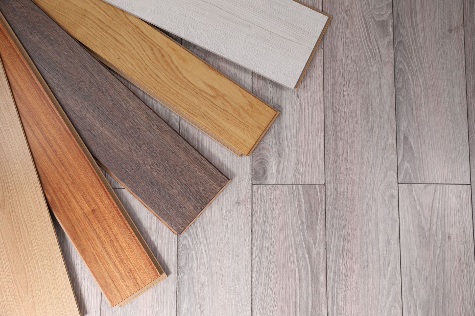 A selection of wooden flooring planks in various shades of brown, tan, and grey, arranged over a light grey wood floor.