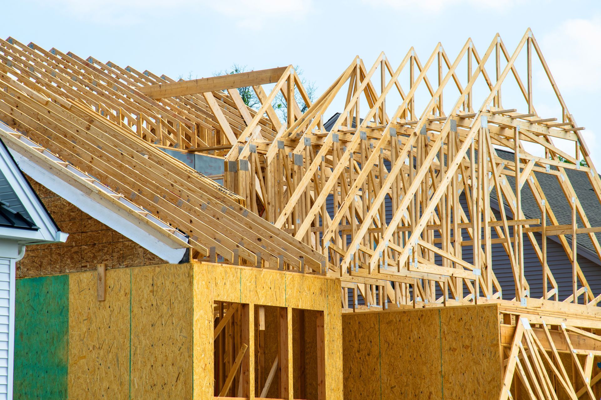 A new house under construction showing wooden wall panels and complex roof truss framing against a blue sky.