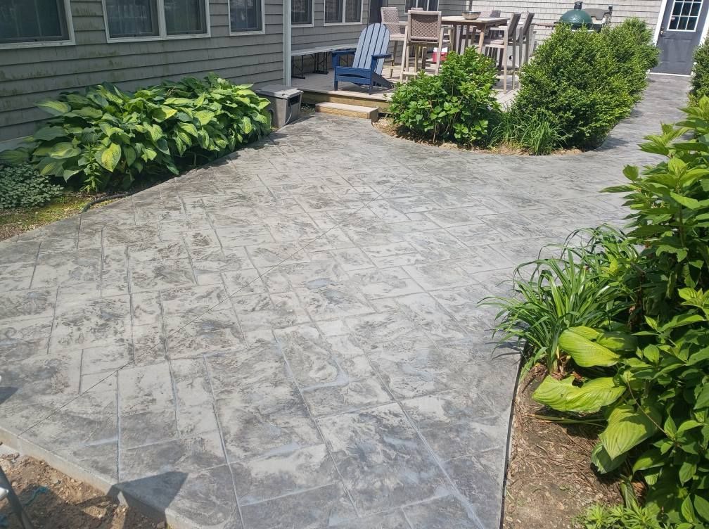 A grey stamped concrete patio area beside a house, framed by green garden plants and a small outdoor seating area.