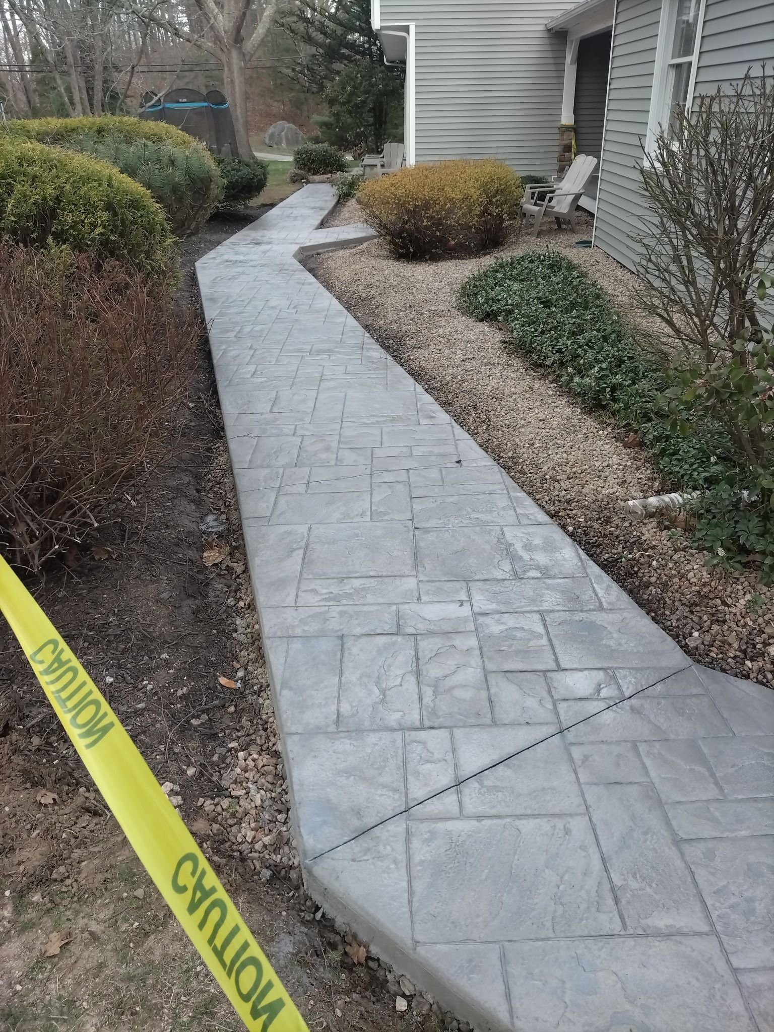 A new gray stamped concrete walkway leads toward a house entrance, bordered by landscaping and caution tape.