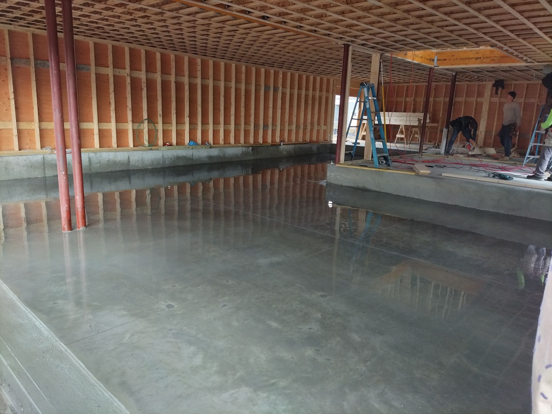 Construction workers finish a smooth concrete floor inside a structure with wooden wall frames and support pillars.