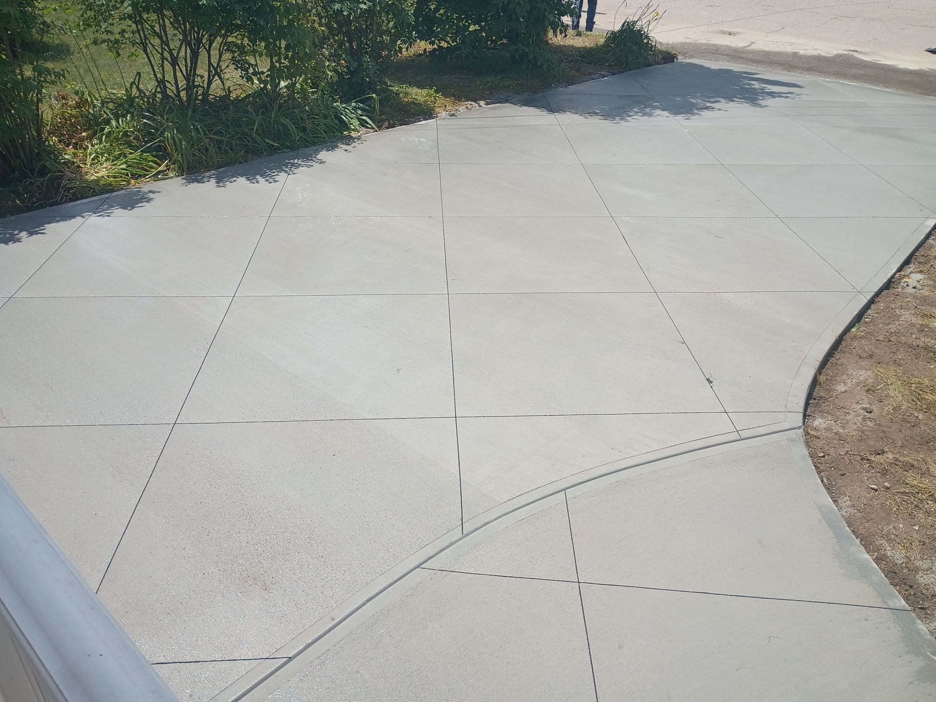 A newly poured concrete driveway featuring a grid of control joints, with green bushes and dirt along the edges.