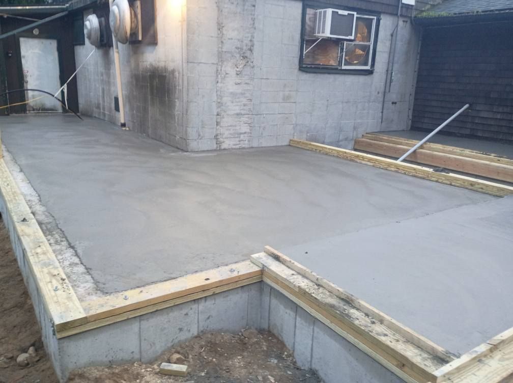 Freshly poured concrete slab with wooden formwork bordering the edge, set against a building foundation.