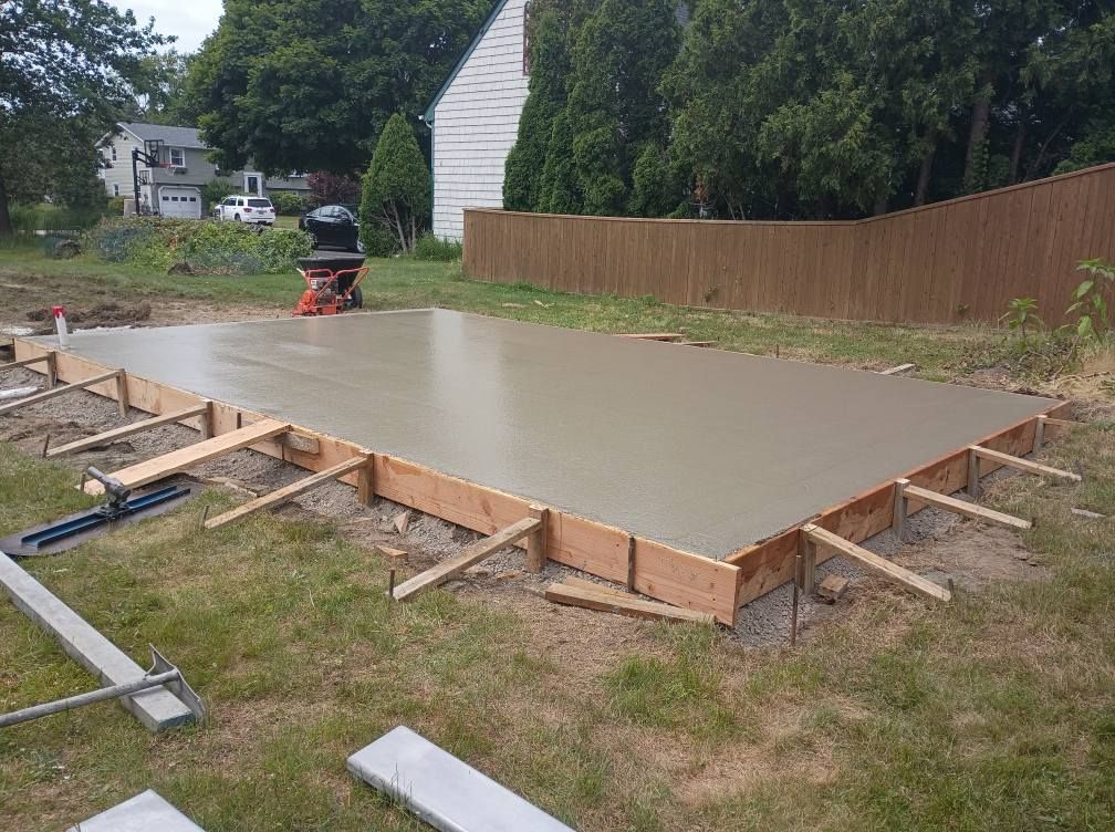 A freshly poured concrete slab in a backyard, enclosed by wooden forms supported by angled stakes on a grassy lawn.