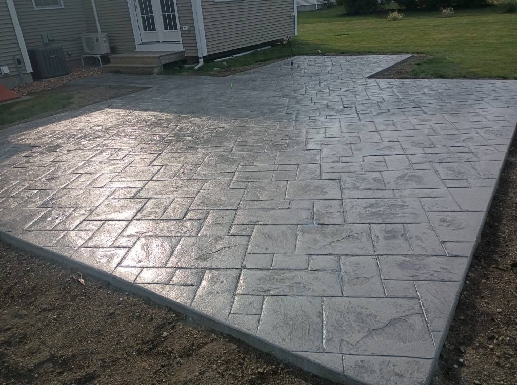 A light gray stamped concrete patio with a rectangular stone pattern set against the side of a house and grassy yard.