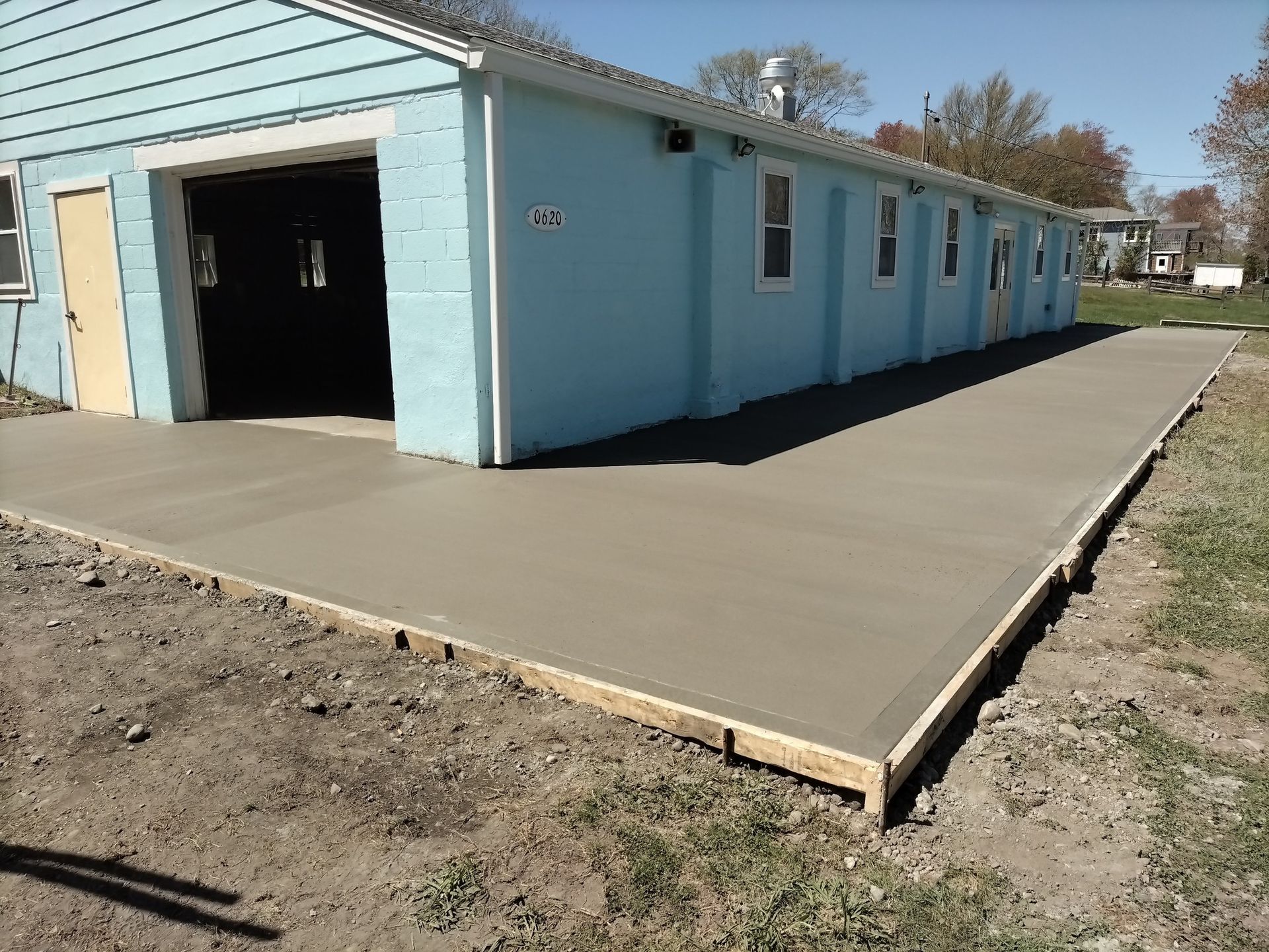 A freshly poured concrete patio runs along the side of a light blue building with a large open doorway.