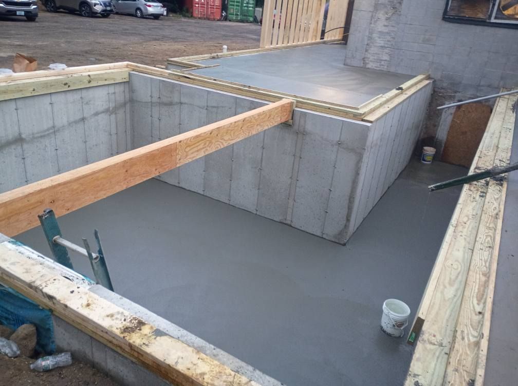 Freshly poured gray concrete floor inside a foundation framed by wooden beams and concrete walls at a construction site.