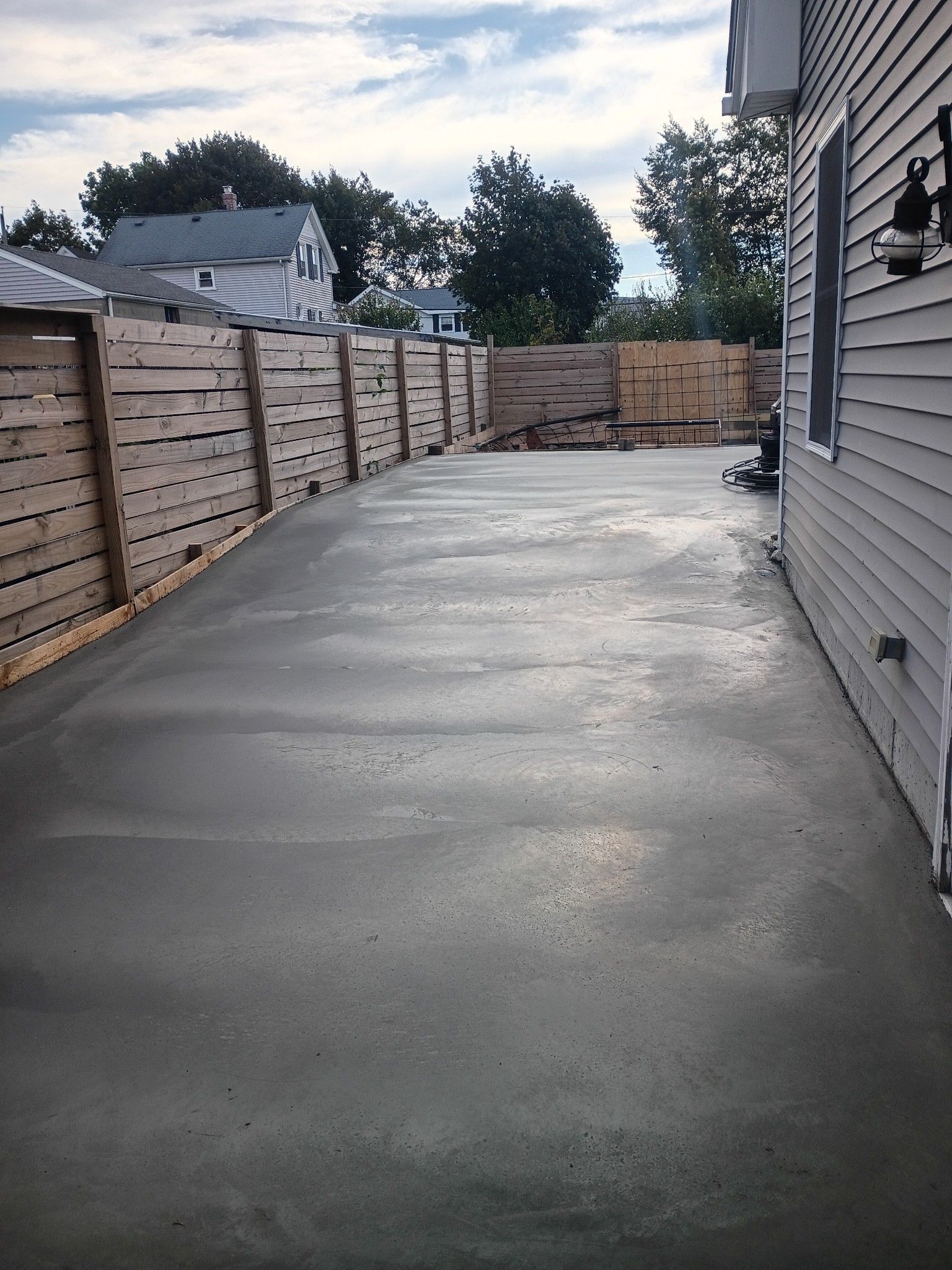 A freshly poured gray concrete driveway next to a light gray house and a wooden horizontal-slat fence.