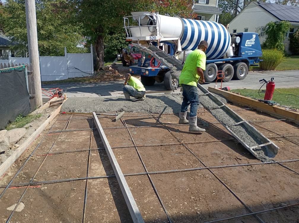 A construction worker in a high-visibility shirt pours concrete from a truck mixer into a framed site with metal rebar.