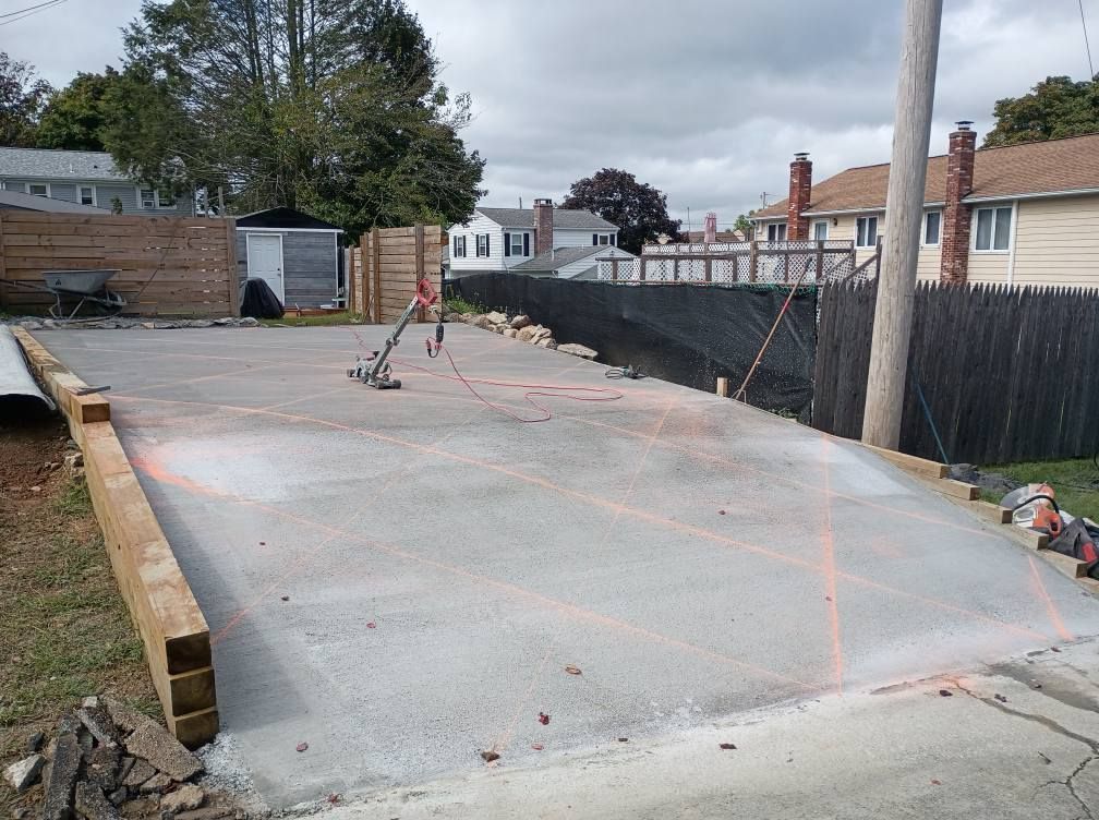 Freshly poured concrete driveway slab with orange guide lines and a tamper tool, surrounded by wooden beams and fences.