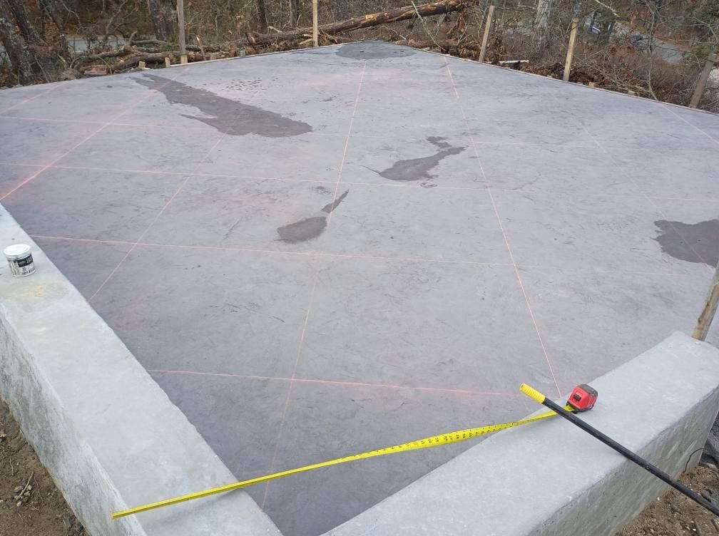 A gray concrete foundation with laser-level lines crossing the surface, viewed from a corner with a measuring tape nearby.
