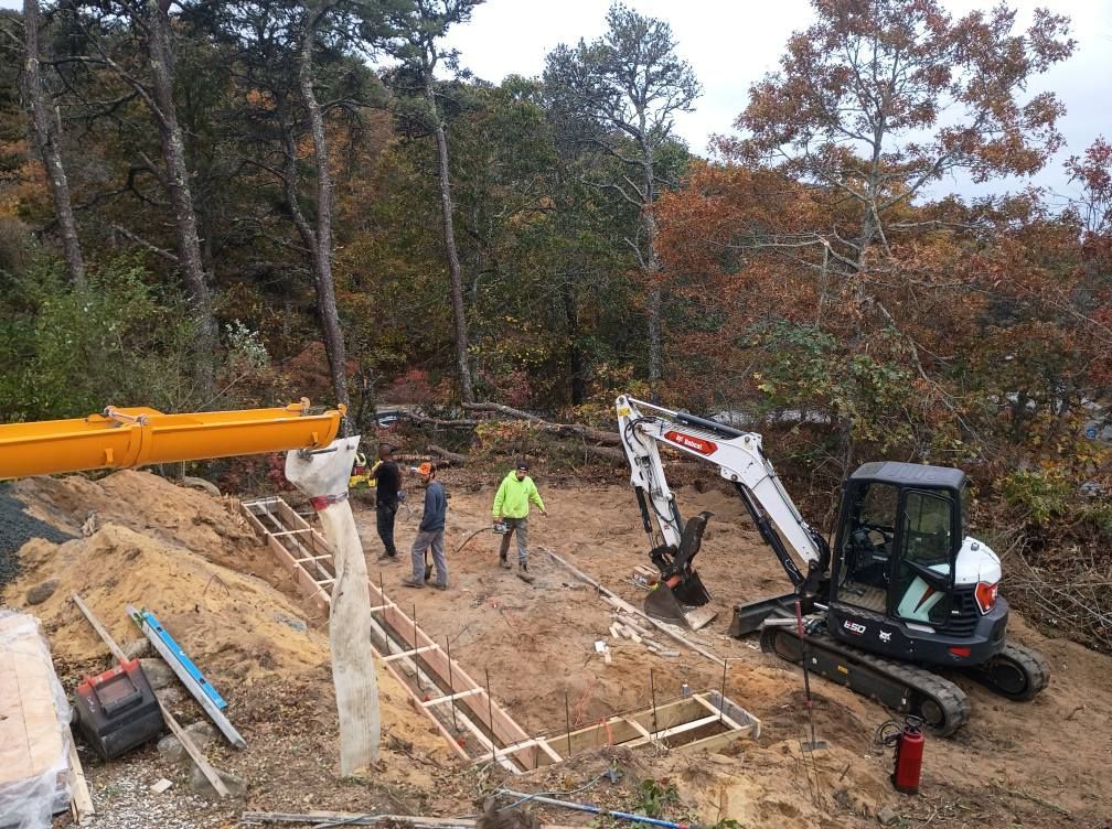 Construction workers prepare a foundation site with a yellow boom crane and a white excavator in a wooded area.
