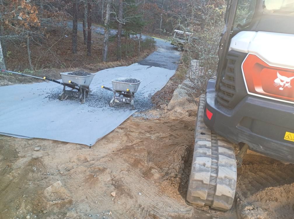 A Bobcat excavator sits on a dirt path next to two wheelbarrows resting on a protective gray ground cover.