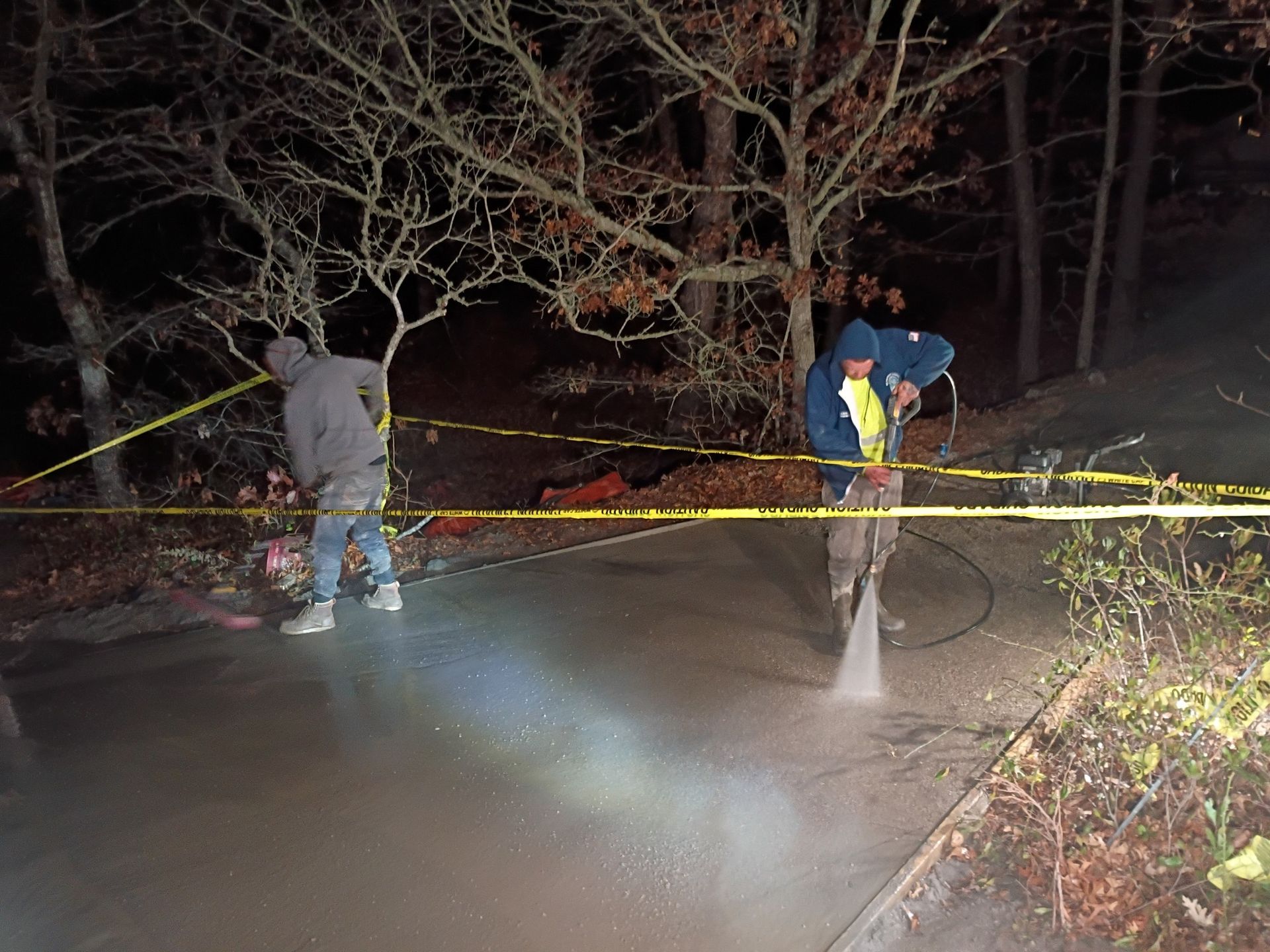 Two people in hoodies use a pressure washer on a concrete driveway at night, surrounded by yellow caution tape.