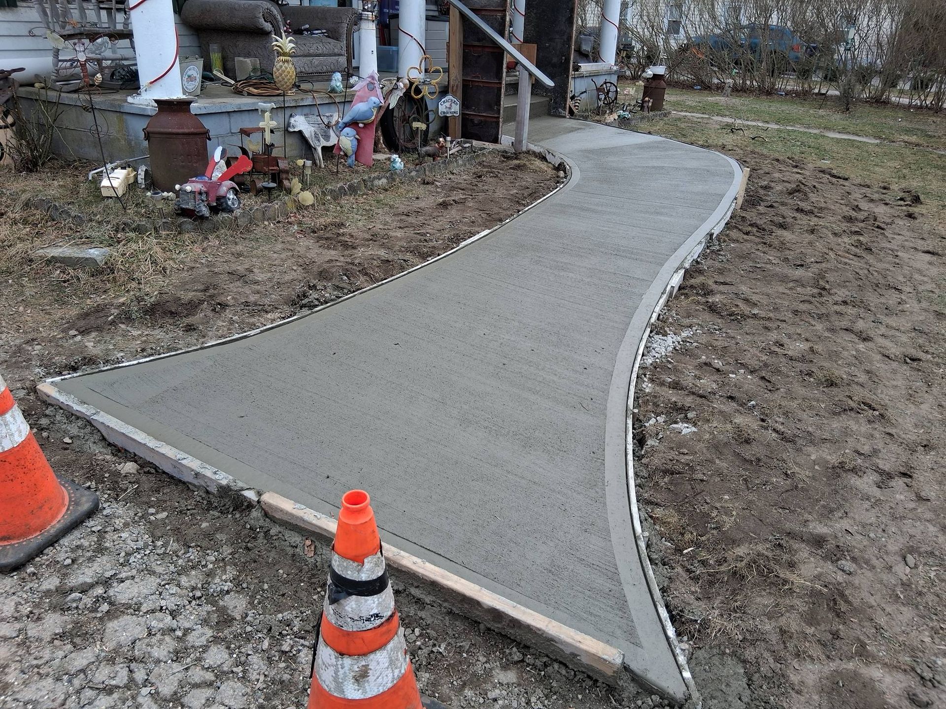 A newly poured concrete walkway curves toward a front porch, flanked by construction cones and bare ground.