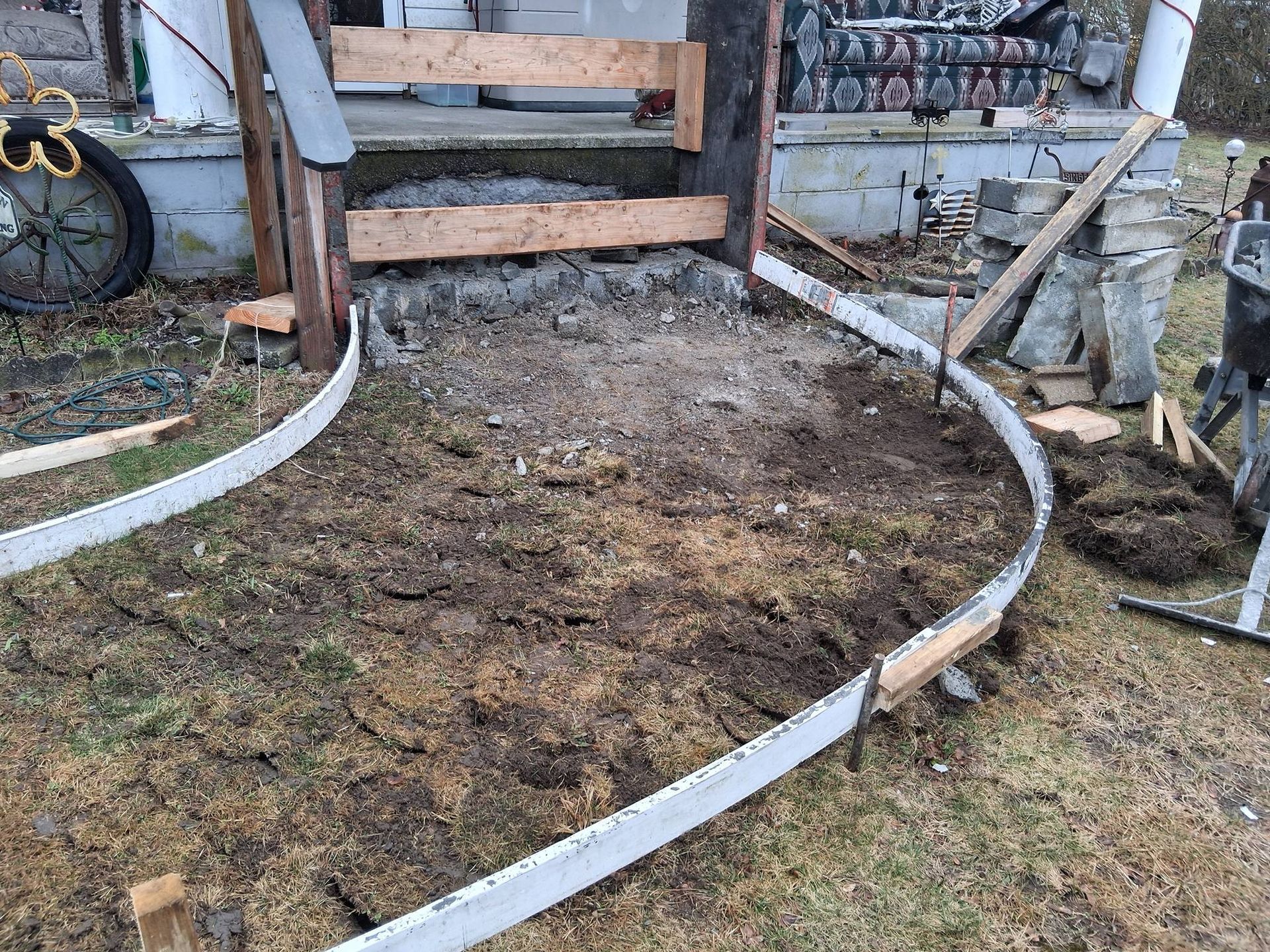 An outdoor construction site showing a curved concrete form set in the ground leading up to a wooden porch staircase.