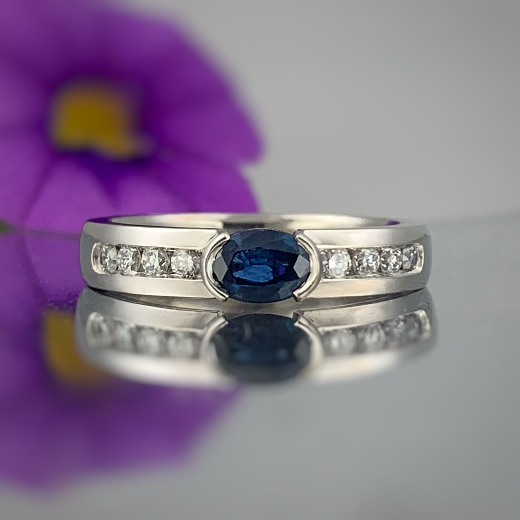 A sapphire and diamond ring is sitting on a table next to a purple flower.
