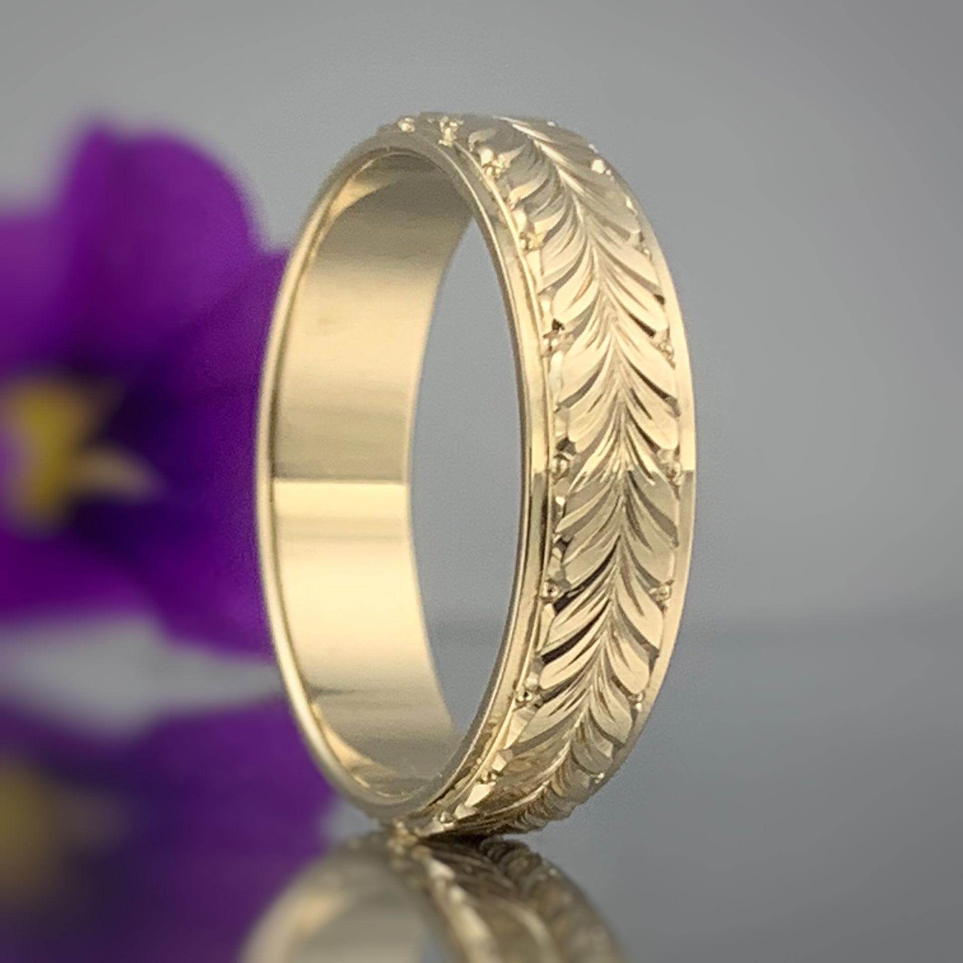 A gold ring with a feather design is sitting on a table next to a purple flower