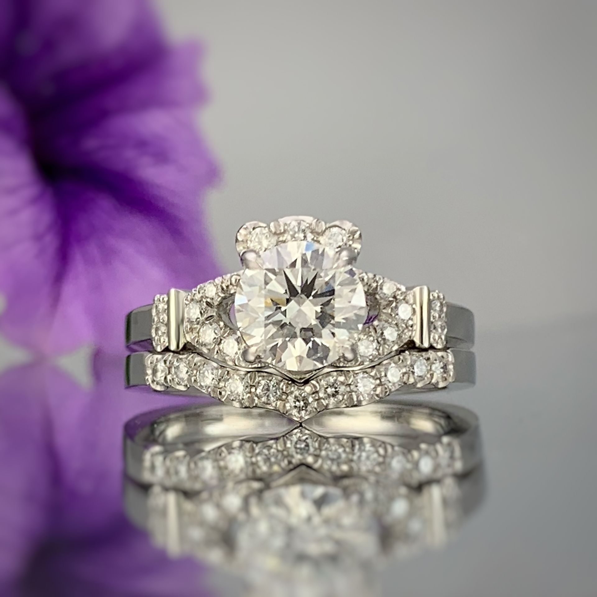 A wedding ring and engagement ring are sitting on a table next to a purple flower.