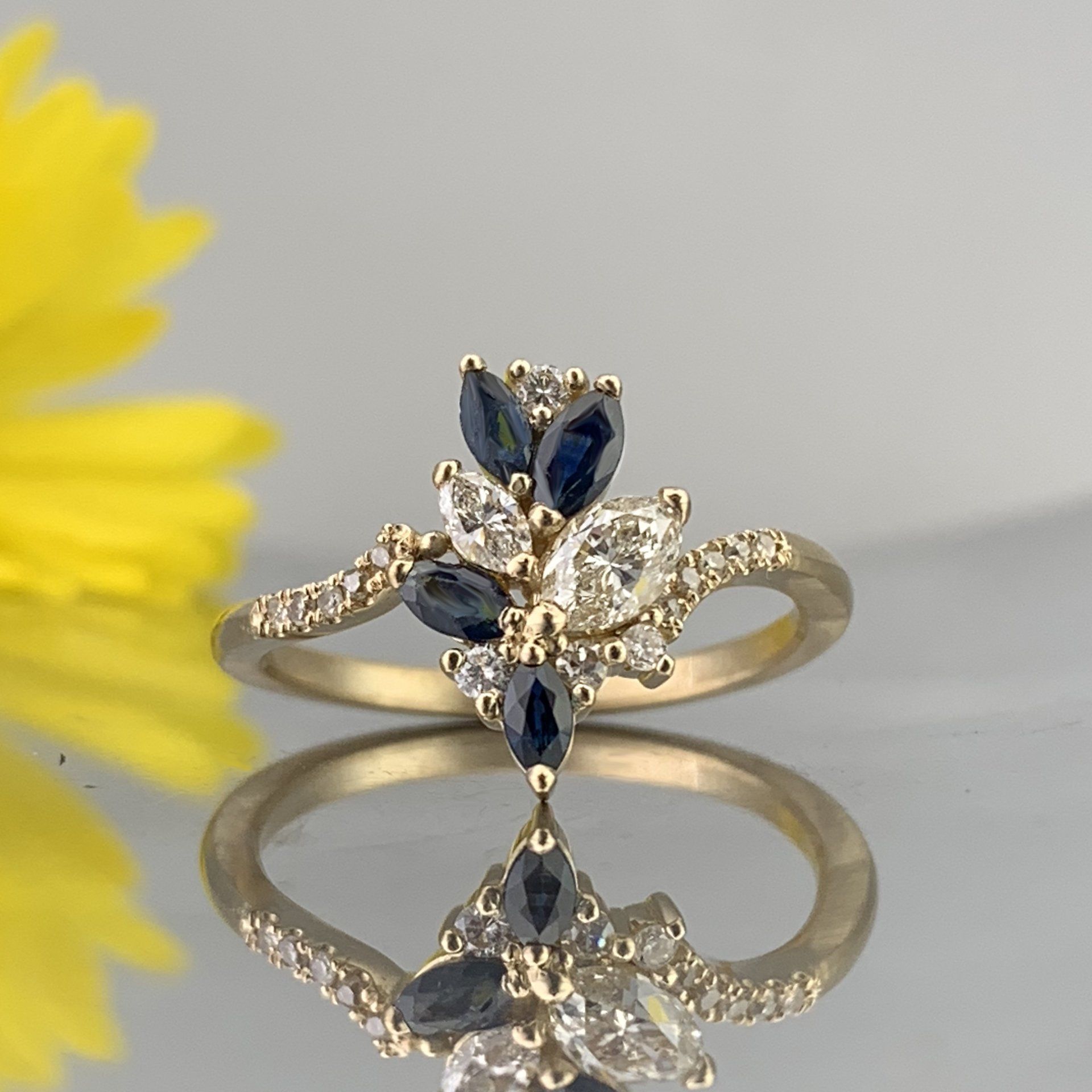 A gold ring with sapphire and diamonds is sitting on a table next to a yellow flower.
