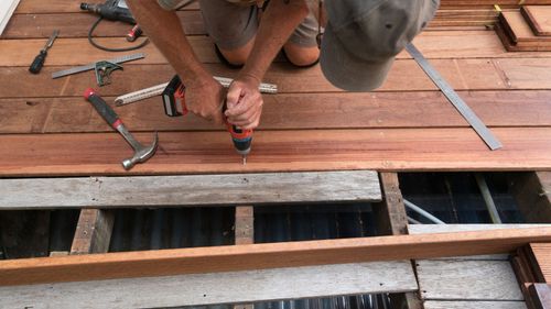 A person kneeling, drilling into planks during deck repair, surrounded by tools.