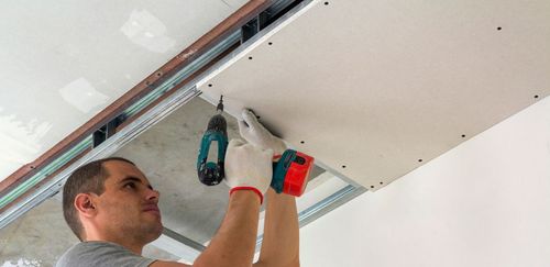 A man in gloves using a drill to install drywall on a ceiling.