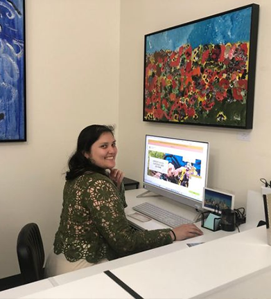 Woman with long, wavy brown hair smiles, leaning against a dark brick wall.