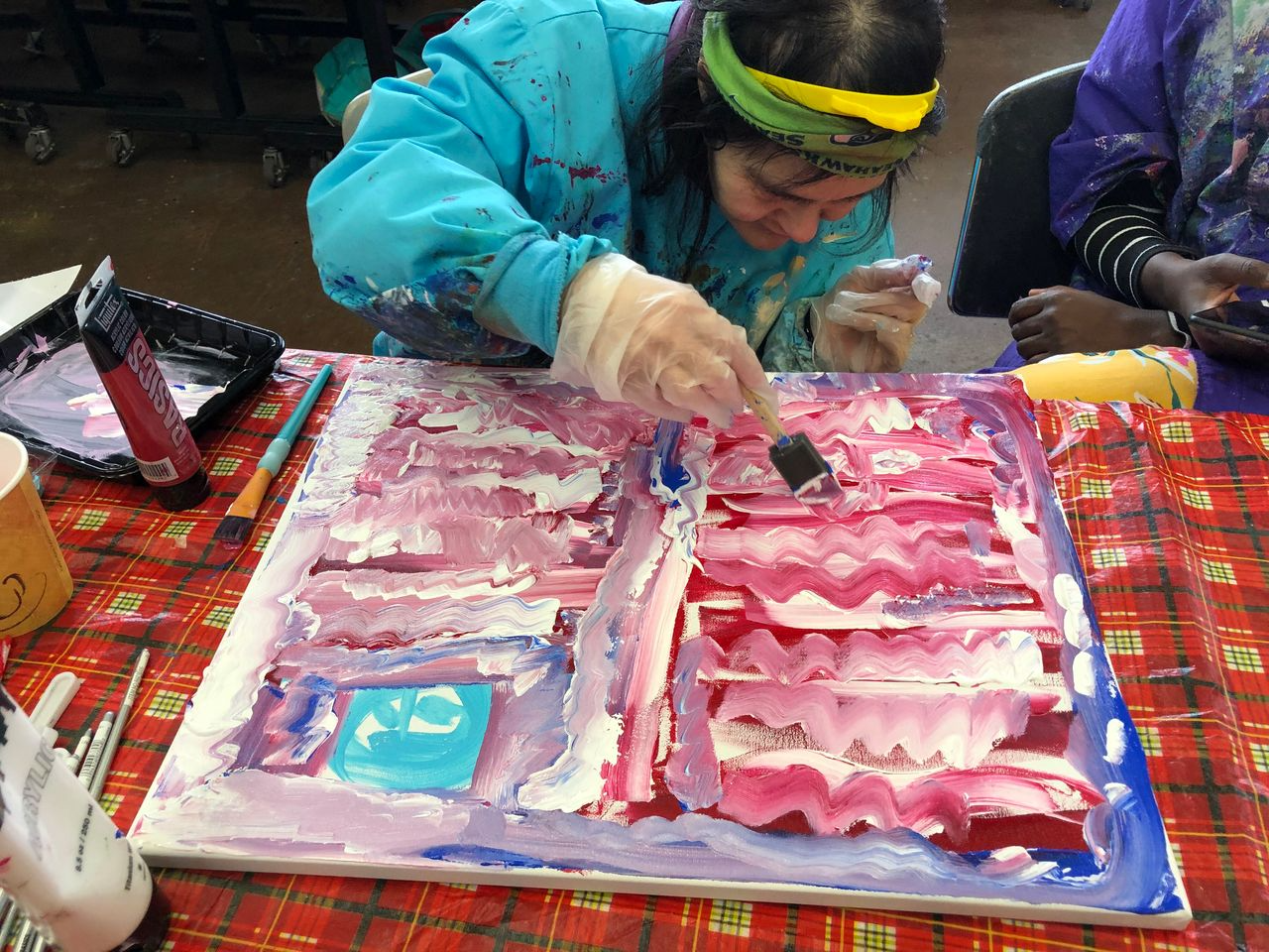 Painting of a red rose in a blue vase on an easel. Other framed artwork hangs on a blue wall.