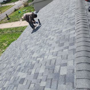 A man is kneeling on top of a roof looking at the shingles.