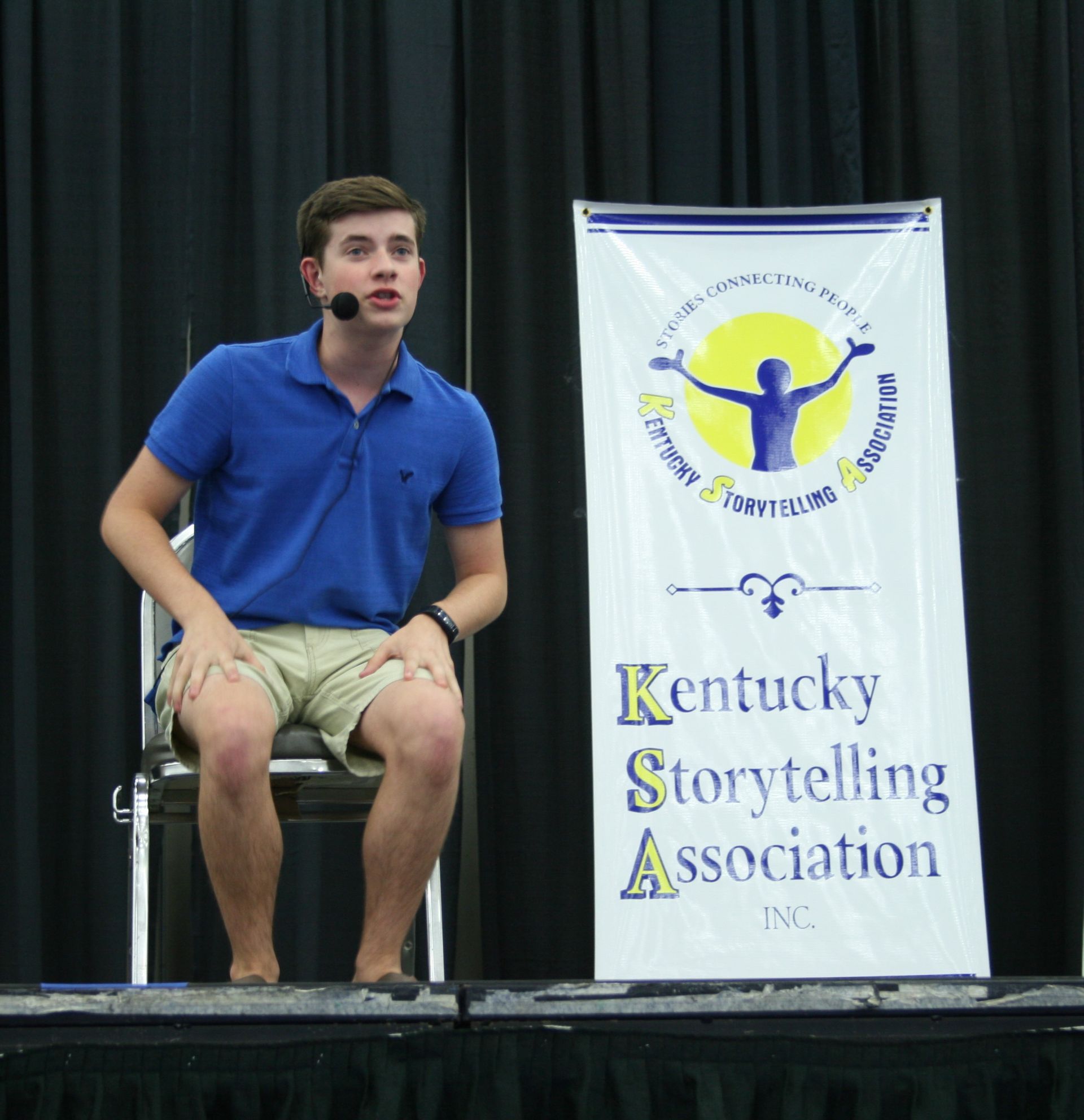 A seated young man on stage with a microphone next to a sign for the Kentucky Storytelling Association.