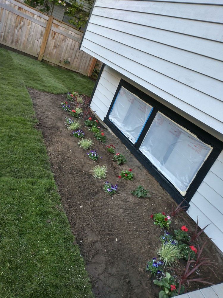A flower bed next to a house with various colorful flowers and green grass.