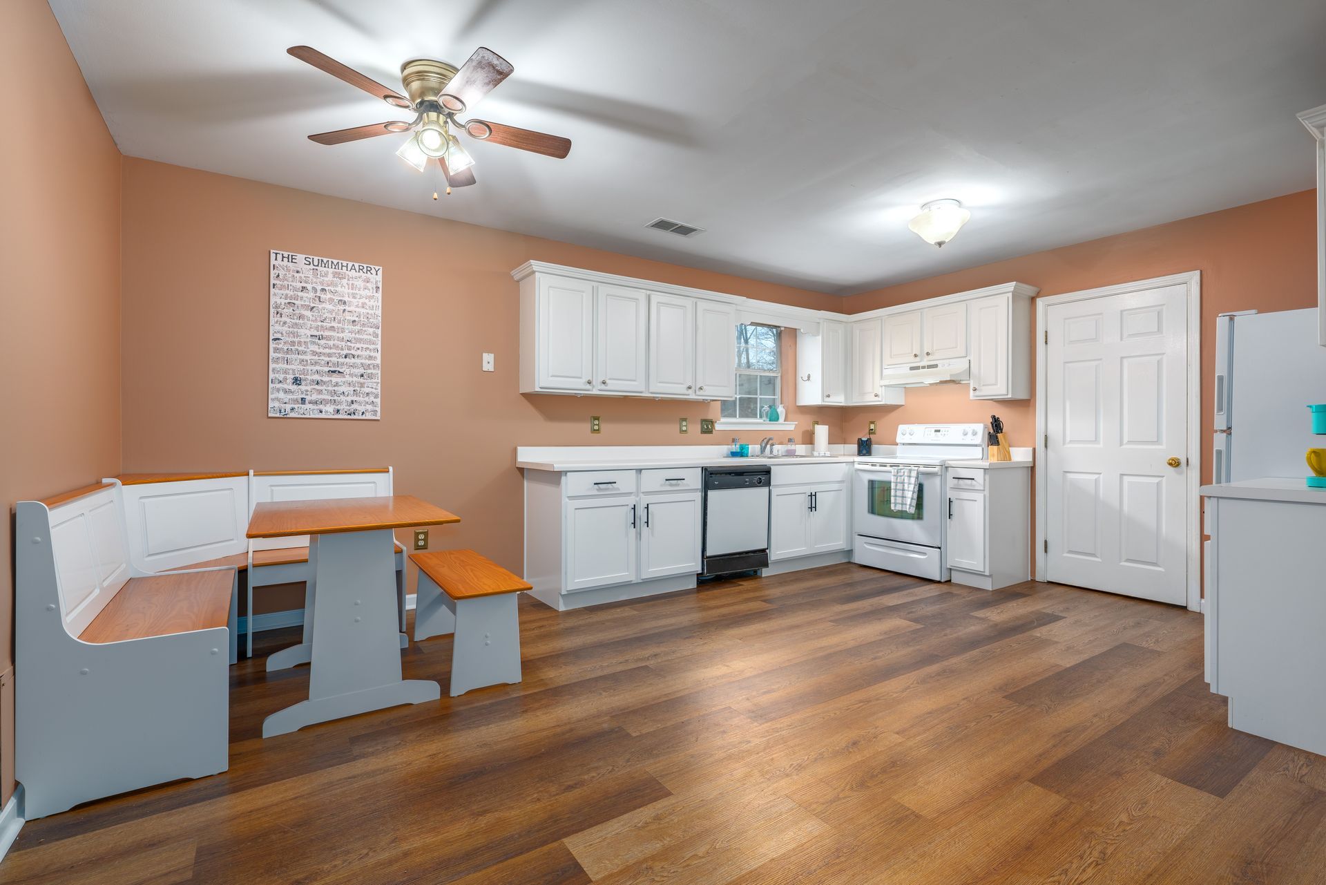 A kitchen with white cabinets , a stove , a refrigerator , a bench and a ceiling fan.