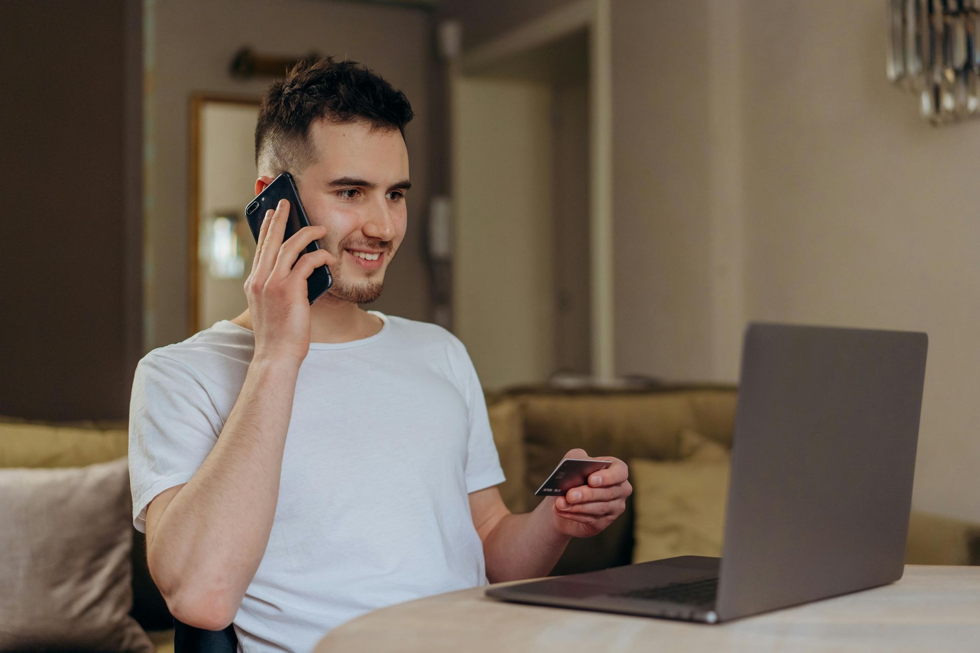 Man on phone, smiling, holding credit card while using a laptop in a living room setting.