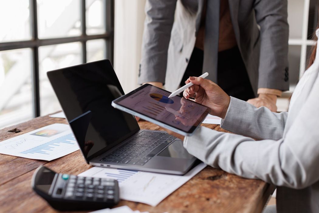 Two professionals in business attire collaborate at a wooden desk with a laptop, tablet, calculator, and financial charts. Two professionals in business attire collaborate at a wooden desk with a laptop, tablet, calculator, and financial charts.