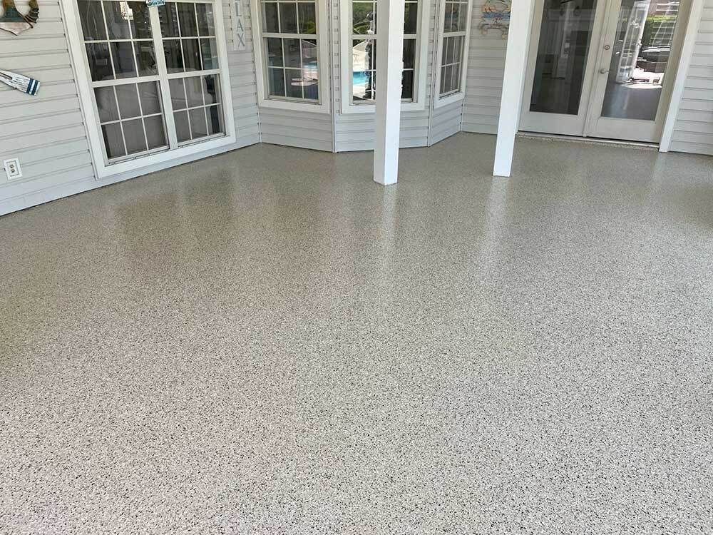 Covered porch with light-colored speckled flooring, windows, columns, and a doorway.