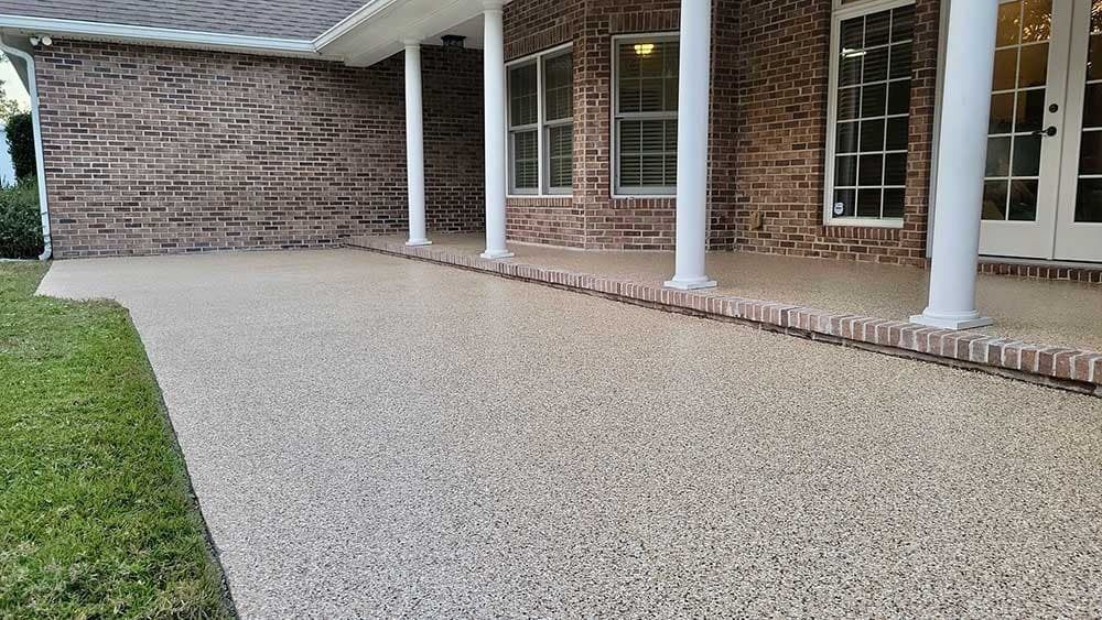 Covered porch with light-colored pebble-textured concrete flooring, brick walls, white columns, and green grass.