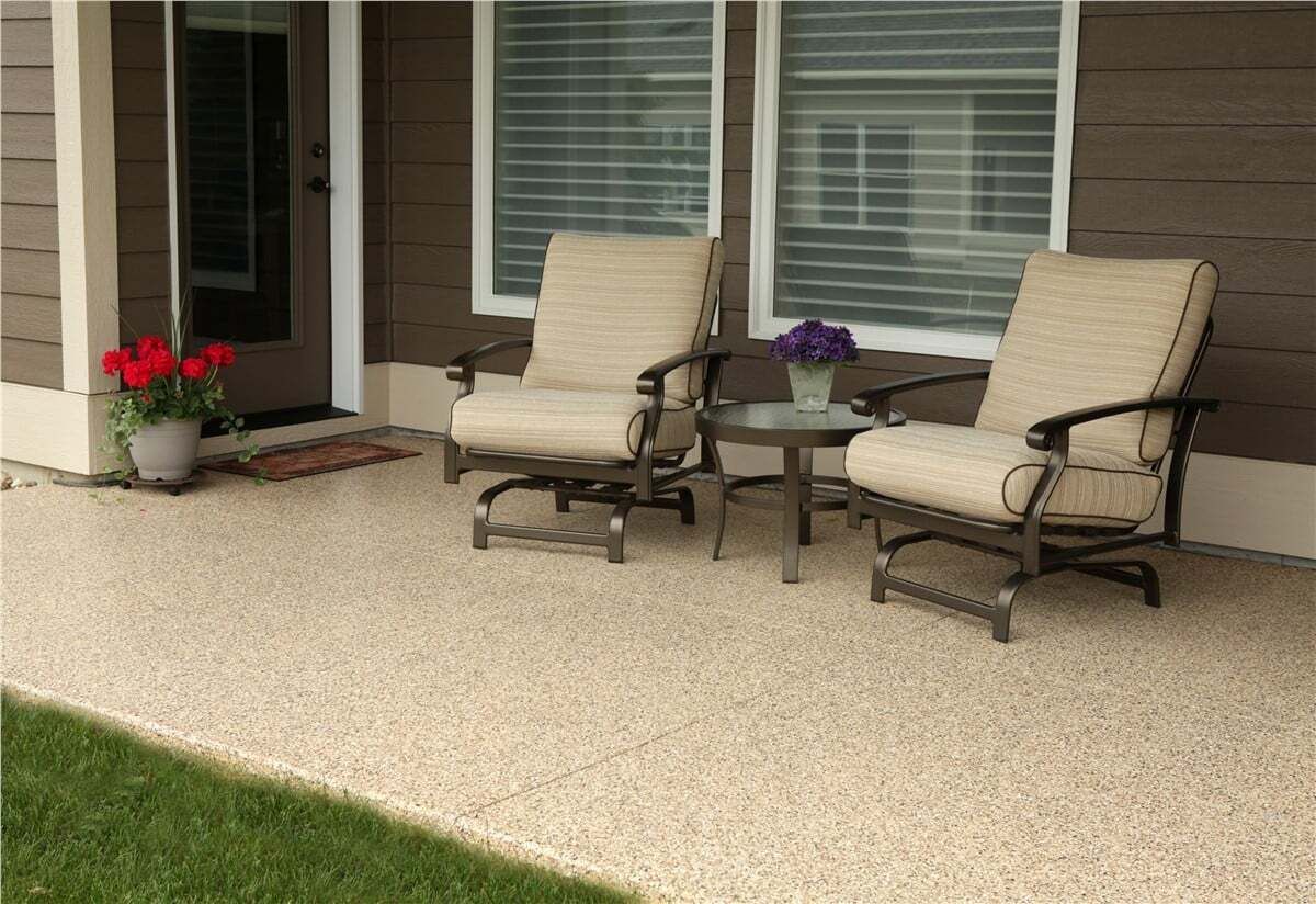 Two beige cushioned chairs on a speckled patio with a small table, door, and potted flowers.