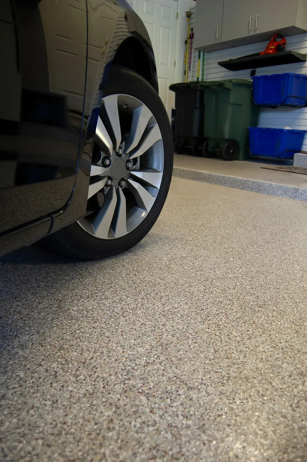 Black car parked in a garage with speckled, grey floor. Garbage bins and storage containers are in the background.
