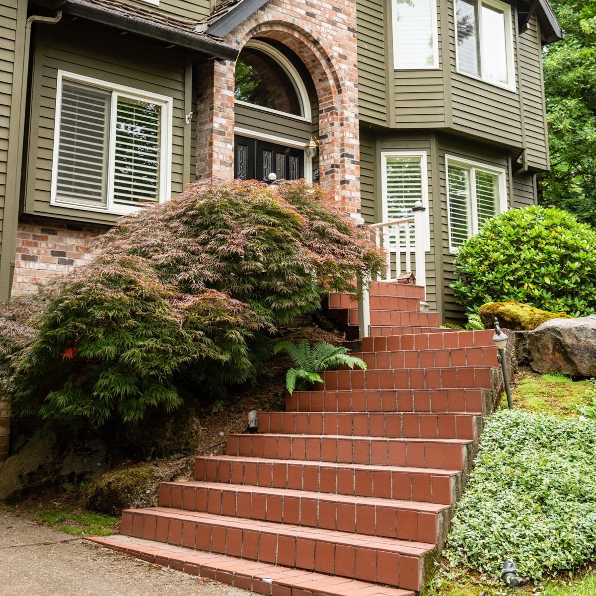 Brick steps lead up to a two-story green house, with a red brick archway and an overgrown bush.