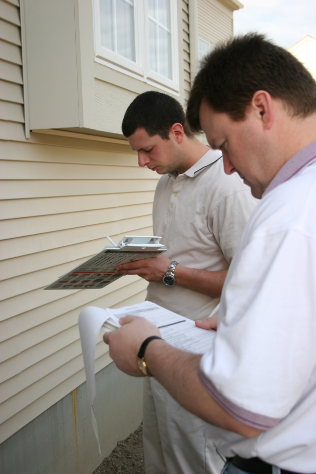 Two men inspecting a house exterior, one holding a clipboard, the other a tablet.